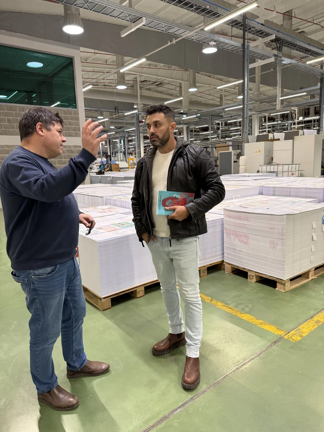 André Braga, com um exemplar do Sem Perdão na mão, à conversa com um funcionário no meio de paletes de papel numa gráfica.